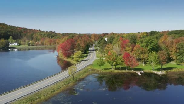 風光明媚な紅葉の森と大規模な湖に沿って航空カントリーロード 晴れた秋の日に湖の海岸で映画的なカラフルな紅葉の木 落下木と道路上のドローンショット メイン州道の旅 — ストック動画