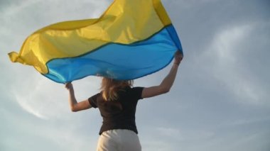 Sarışın bir kadın Ukrayna bayrağını kafasının üstünde tutuyor ve geriye doğru koşuyor. Zafer işareti olarak aktif olarak koşuyor. Ağır çekim. Yüksek kaliteli FullHD görüntüler