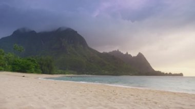 Yavaş çekim nefes kesici sinematik doğa manzarası berrak boş plaj, yağmur ormanları dağ parkında şelale. Yüksek yeşil dağlar üzerinde dramatik gri bulutları olan tropik plaj manzarası