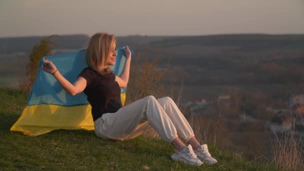Sarışın Bir Kadın Tepede Oturmuş Günbatımını Izliyor Ukrayna Bayrağıyla Sarılmış — Stok video
