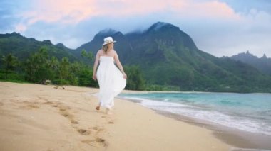 Hawaii 'de yaz tatili adasında şık bir gezgin. Gün batımında güzel beyaz sallanan elbiseli bir kadın. Gün batımında kumsalda çıplak ayakla yürüyen kadın kumsalda ayak izleri bırakıyor.
