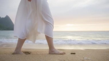 Ufkun yukarısındaki altın gün batımında kumsalda çıplak ayakla yürüyen beyaz boho elbiseli kadın kumsalda ayak izleri bırakıyor. Hawaii 'deki Kauaii adasında yaz tatilindeki kadın turist.
