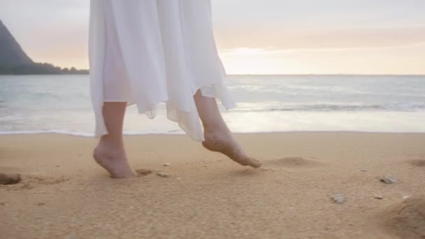Zpomalená žena v bílých boho šatech kráčí po špičkách po pláži při zlatém západu slunce nad obzorem a zanechává stopy v písku. Bosá žena turista na letní dovolenou na ostrově Maui Hawaii USA cestování
