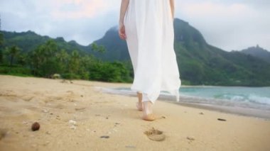 Hawaii 'deki Kauai Adası' nda düğününü kutlayan mutlu bir kadın. Güzel beyaz sallanan elbiseli bir kadın kumların üzerinde ayak izleri bırakıyor. Gün batımında çıplak ayakla yürüyen gelinlik giymiş yavaş çekim gelini.