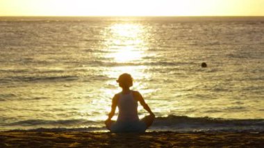 Tanımlanamayan genç yogacı kadının tropikal plajda altın rengi günbatımı ya da gün doğumu ile Lotus pozisyonunda oturması. Hawaii 'de yoga yapan bir kadının arka görüntüsü.