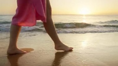 Yaz tatilindeki kadın turist gün batımında Hawaii adasında okyanus kıyısında altın sıvıyla yürüyor. Yavaş çekim kadın ayakları, altın gün batımında kumsalda yalın ayak yürüyor ıslak kumda ayak izleri bırakıyor.