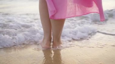 İnce kadın bacakları ve ayakları birbirine yakın kumlu sahilde deniz dalgalarında duran bir kadın. Deniz kenarında sörf yapan pembe elbiseli güzel bir kadın. Su ve köpük damlaları yavaş çekimde. Okyanusta yıkandıktan sonra kız.