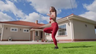 Dişi sporcu, yemyeşil çimlerde kardiyo çalışması yapıyor. Dışarıda kırmızı eşofmanlı bir kadın koşuyor. Yüksek kalite 4k görüntü