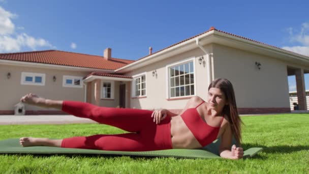 年轻的高加索女人做侧腿举重 后面有漂亮的房子 女运动员躺在瑜伽垫上 同时训练她的髋关节和光环 高质量的4K镜头 — 图库视频影像