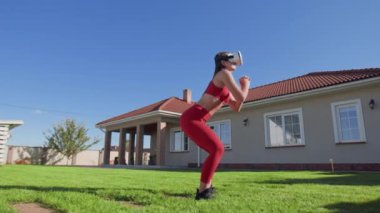 Dışarıda güzel bir evi olan sanal gerçeklik kulaklığı kullanan genç bir kadın. Ev spor salonunda çömelme hareketleri yapan bir bayan sporcu. Yüksek kalite 4k görüntü