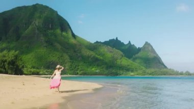 Yavaş çekim mutlu kadın romantik plaj elbisesi giyer. Sinematik okyanus plajında koşar. Gün doğumunda hareketli yeşil dağ zirveleri vardır. Mutlu ve hayattan sarhoş, Hawaii 'nin güzel manzarasının tadını çıkarıyor.