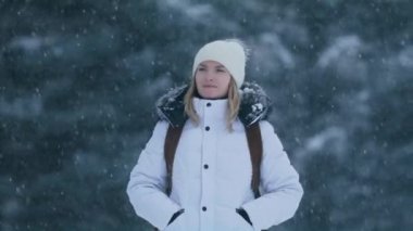 Sırt çantalı, güzel, sarışın, gülümseyen bir kadının yavaş hareket eden yüzü. Kar yağışlı kış gününde yeşil ormana bakan muhteşem bir kızın portresi. Bulanık yeşil ağaçlarda güzel bir gezgin.