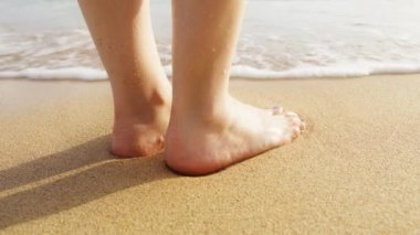 Yaz tatilinde, okyanus dalgalarında kıyıda duran yakın bir kadın. Ağır çekimde sahne arkası. Islak sahil yüzeyinde güzel altın güneş ışığı. Kumsalda çıplak ayaklı bir kadın.