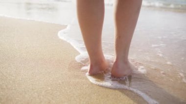 Woman feet walking barefoot on sandy beach of sea. Scenic summer background in slow motion. Beautiful golden sunlight reflecting from the wet beach surface.Woman standing in ocean waves on sandy beach