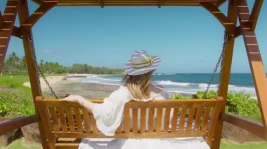 Ağır çekimde gevşemiş, romantik beyaz şapkalı, tropikal çiçekli, okyanus esintisiyle sallanan sarı saçlı kadın. Sinematik ada plajı, rüya adası seyahati, romantik yaz tatili.