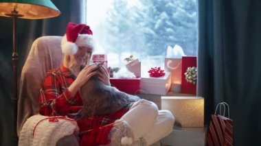 Hala kadınların elindeki kediciğin kırmızı kamera görüntüsü. Gri tüylü sevimli hayvanını okşayan bir kadın. Kışın iç mekanda, Noel günü, pencerede Noel hediyeleriyle oturan bir kadın.