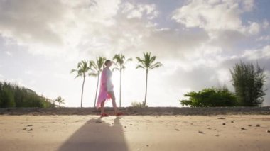 Woman in pink dress enjoys strolling along the beach with sunny sky in the background. Female tourist walks around isolated island with tropical vegetation. High quality 4k footage