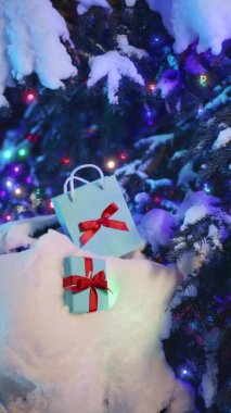 Sihirli kış arka planının dikey mobil içeriği. Mutlu Noel arifeleri. Sürpriz mavi hediye çantası, kırmızı saten fiyonklu hediye kutusu. Karlı köknar ağacının dalında güzel hediyeler.