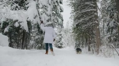 Cheerful woman in white puffer coat and pom-pom hat having fun and playing with dog in winter forest. First snow in magical mountain forest. Slow motion winter wonderland shot on RED camera in 6K