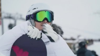 Beyaz kasklı, aktif bir kadının portresi ve kar yamacında mükemmel bir kış gününde mor snowboard 'a yaslanan renkli Google' lar. Arka planda kayak pistleri 6K