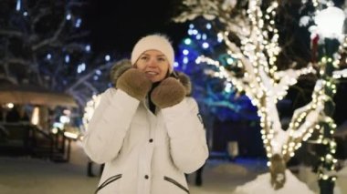 Gülümseyen güzel bir kadının portresi bulanık arka planda şehir parkında manzaralı aydınlatma ile Noel gecesinden keyif alıyor. Tatil tatili 6K. Kasabanın önünden geçen kışlık ceketli olumlu bir kadın.