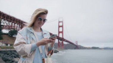 Golden Gate 'in dışındaki akıllı telefon cihazıyla cep telefonu tutan mutlu beyaz kadın. Gülümseyen genç kadın blogcu yeni sosyal medya aboneliği, internetten satın alma, uygulamalardan ürün sipariş etme