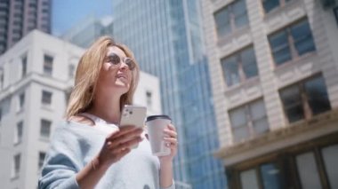 Kentin hareketli arka planında elinde fincanlı kahve ve akıllı telefonla duran yakın plan neşeli kız. Dışarıda cep telefonunu tutan gülümseyen kadın. Dışarıda cep telefonuyla yazan mutlu hipster kadın portresi.