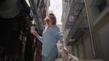 Alışveriş torbası ve bir fincan kahveyle yürüyen genç ve mutlu bir kadının arka görüntüsü. Kameraya gülümseyen ve saçlarını dalgalandırarak flört eden bir kız. Güzel bir kadın Dar şehir sokağında uzaklara gidiyor