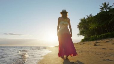 Beautiful summer vacation on paradise island, slow motion young woman walking towards the golden sunrise with lens flare from cinematic sun rays. Traveler on amazing pure ocean beach at scenic sunset