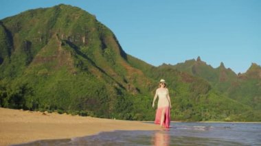 RED camera woman on island trip or adventure. Young traveller in long pink dress or travel blogger walk by beautiful ocean beach on mountain landscape. Perfect summer vacation, concept travels Hawaii
