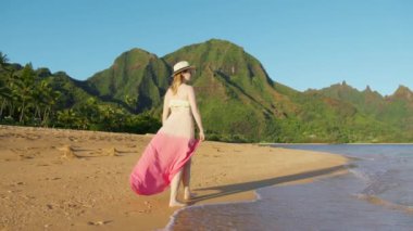 Happy romantic traveler woman enjoying relaxing sunset or sunrise walk on Hawaii beach. Slow motion back view of tourist female having fun walking down beach to beautiful tropical green mountain park