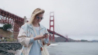 Arka planda, dünyaca ünlü Golden Gate Köprüsü yakınlarında yürüyen turist kadın. Akıllı telefondan mesaj atan genç sarışın kadın. RED 'de yavaş çekim. San Francisco 'da akıllı telefonlarda uygulama kullanan bir kadın.
