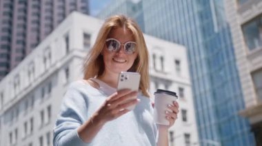Güzel bir kadın ayakta duruyor, büyük şehir caddesinde akıllı telefon kullanıyor. Bokeh arka planındaki gökdelen ofis binalarında. Gülümseyen güzel bir kadın cep telefonu kullanıyor. Kızıl Kamera 'ya yakın çekimde sinematik çekim