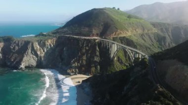 Cinematic aerial shot SUV cars driving on sunny clean road highway to mountains. Summer vacation dream travel, road trip by cinematic nature landscape at Big Sur with Bixby arch bridge on West Coast
