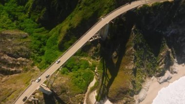 Soaring over long stretch of ocean view highway in the middle of a lush green mountain canyon with rolling hills and steep rocky cliffs raising high above powerful ocean with waves crashing at beach