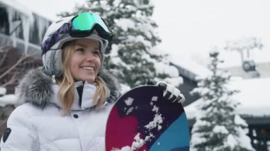 Portrait of gorgeous blonde haired caucasian cheerful woman with pretty smile, standing with snowboard in modern ski resort village, wearing sports helmet winter jacket. Happy young woman enjoys life