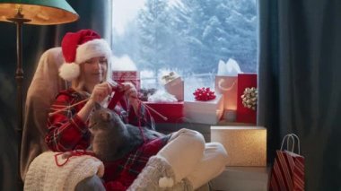 Happy young woman relaxing in cozy home atmosphere with cute cat sitting on her lap and knitting red needles. Woman in Santa hat having fun enjoying handcraft hobby concept at home on winter holidays