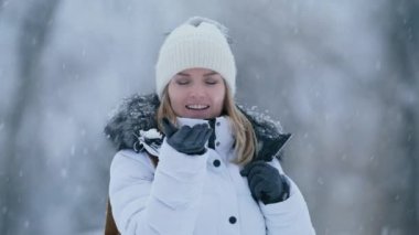 Carefree attractive young woman trying to catch snowflake on the palm of hand in grey gloves. Woman excited by fresh snow and cold winter day with forest on blurred background, shot on RED camera 6K