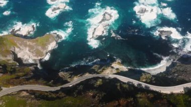 Aerial top view of waves break on rocks in blue ocean. Sea waves beautiful shore aerial view drone 4K shot. Birds eye view of ocean waves crashing at stone rock cliffs from above Big Sur scenic road