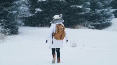 Back view of tourist with backpack hiking by snowy forest on winter day. Young active woman in white coat and beanie hat enjoying winter vacation in wonderland. Slow motion 6K footage people outdoors