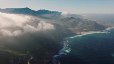 Sunshine hit mountain peaks. Drone flying through clouds. Aerial drone beautiful mountain coast landscape with deep blue Pacific ocean views. Sun rays breaking through white clouds to the rocky shore