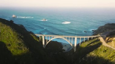 Cinematic ocean waves at sunrise sky on sunny summer morning. West coast road trip by North America, eco nature travel 4K drone. Beautiful scenic arch architecture bridge with blue ocean on background
