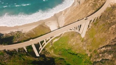 Top down view car driving along scenic arch architecture Bixby bridge above blue ocean waves. Cinematic ocean waves on sunny summer day. West coast road trip, eco nature travel, 4K drone flight USA
