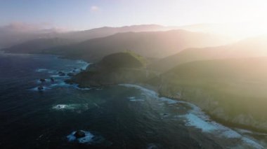 Scenic mountains covered by sunny glowing cloud of ocean breeze. Cinematic aerial nature background. Summer vacation dream travel, road trip by nature landscape at Big Sur on West Coast California USA