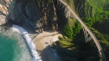 Aerial top down drone shot above scenic bridge on mountain road in Big Sur nature park, California USA. Sunny sunrise on summer vacation day. Car driving by high arch bridge at beautiful Pacific coast