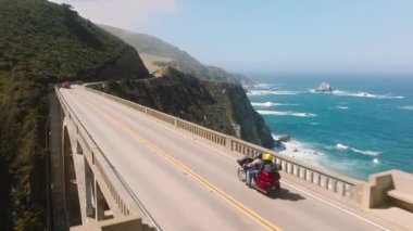 Couple riding on vintage motorcycle by beautiful Big Sur coast with cinematic landscape. Extreme vacation concept, motorbikers on motorcycle rides scenic mountain road. Freedom, life enjoyment aerial