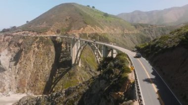 Closeup shot above vehicles roadtripping along Californian coastline. Aerial footage of car crossing the Bixby Canyon Bridge across the mountains. High quality 4k footage