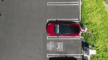 Aerial view of red car charging in green business park. 4K electric, zero pollution cars on green energy concept on modern city parking lot. Alternative energy for ecological cars, zoom out drone 4K
