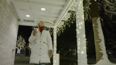 Woman drinks tea a cup while walking by terrace at town village on winter snowy night. Christmas atmosphere, magic feeling, holiday decorations, miracle expectation. Winter holiday vacation, 6K Video
