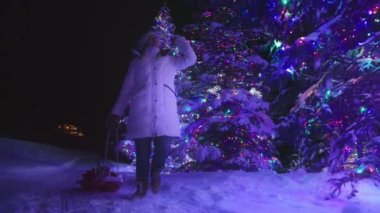 Slow motion Christmas gifts delivery on glowing colorful winter wonderland forest background 6K. Excited young woman in pom pom hat walking by winter wonderland with sleighs full of Christmas presents
